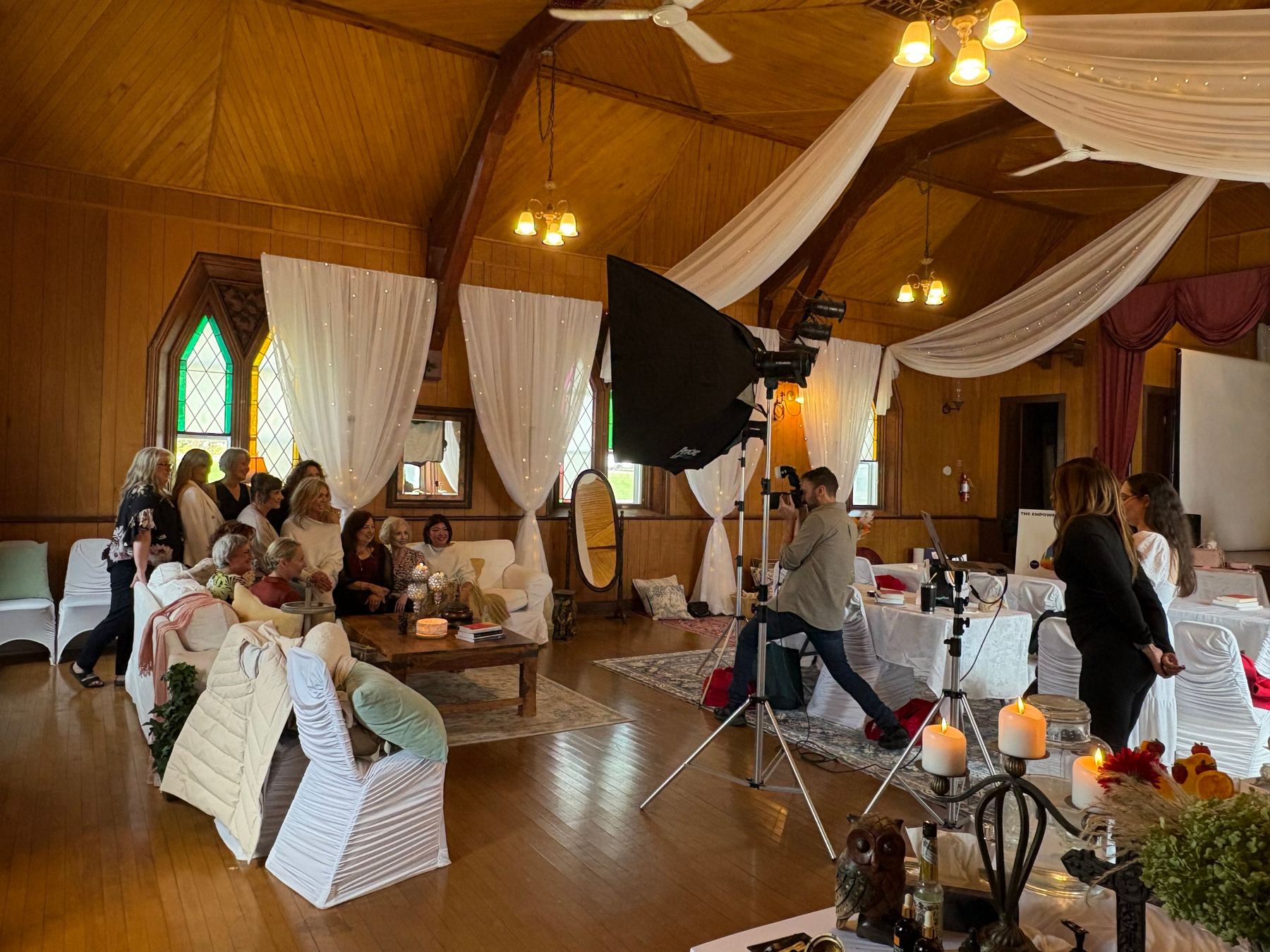 The women of the first gathering on white sofas beneath stained glass while a photographer, softbox lights and crew work around them.
